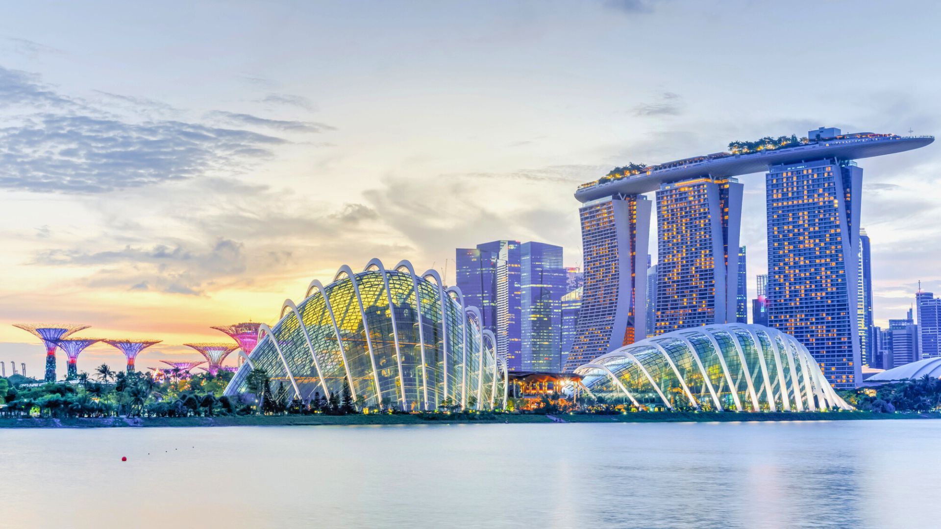 Singapore skyline at dusk featuring Marina Bay Sands and Gardens by the Bay domes, reflected in the water.