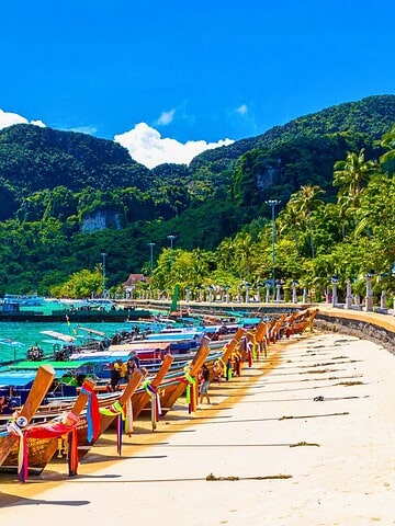 Colorful longtail boats lined up on a sandy beach with turquoise water and lush green mountains in the background.