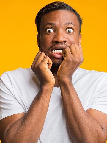 Man in a white t-shirt looking scared, biting his nails with wide eyes, standing against a yellow background.