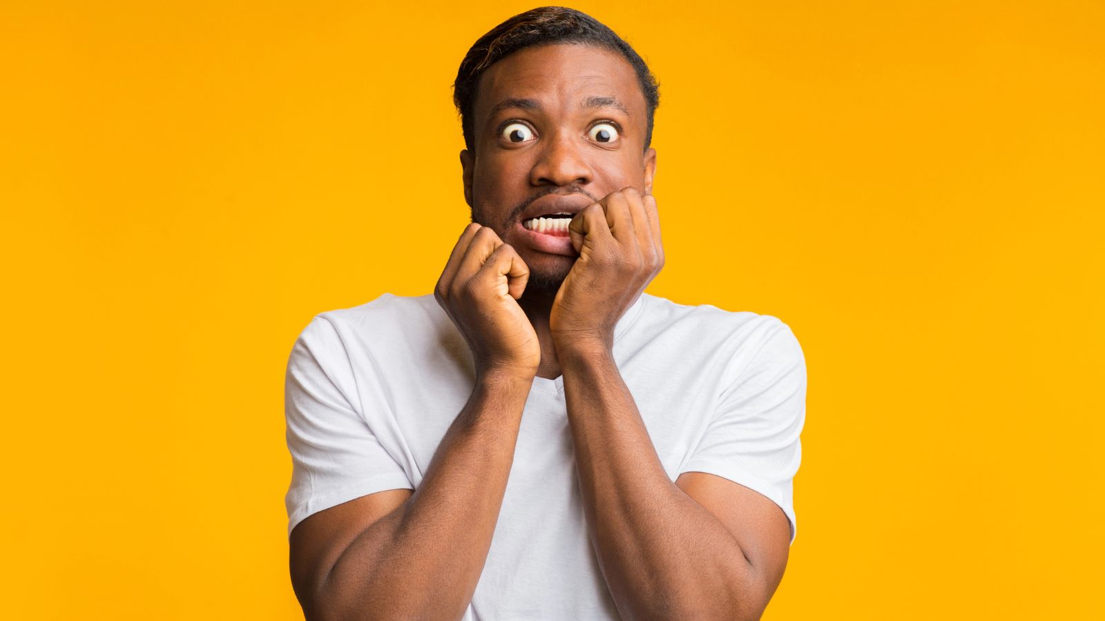 Man in a white t-shirt looking scared, biting his nails with wide eyes, standing against a yellow background.