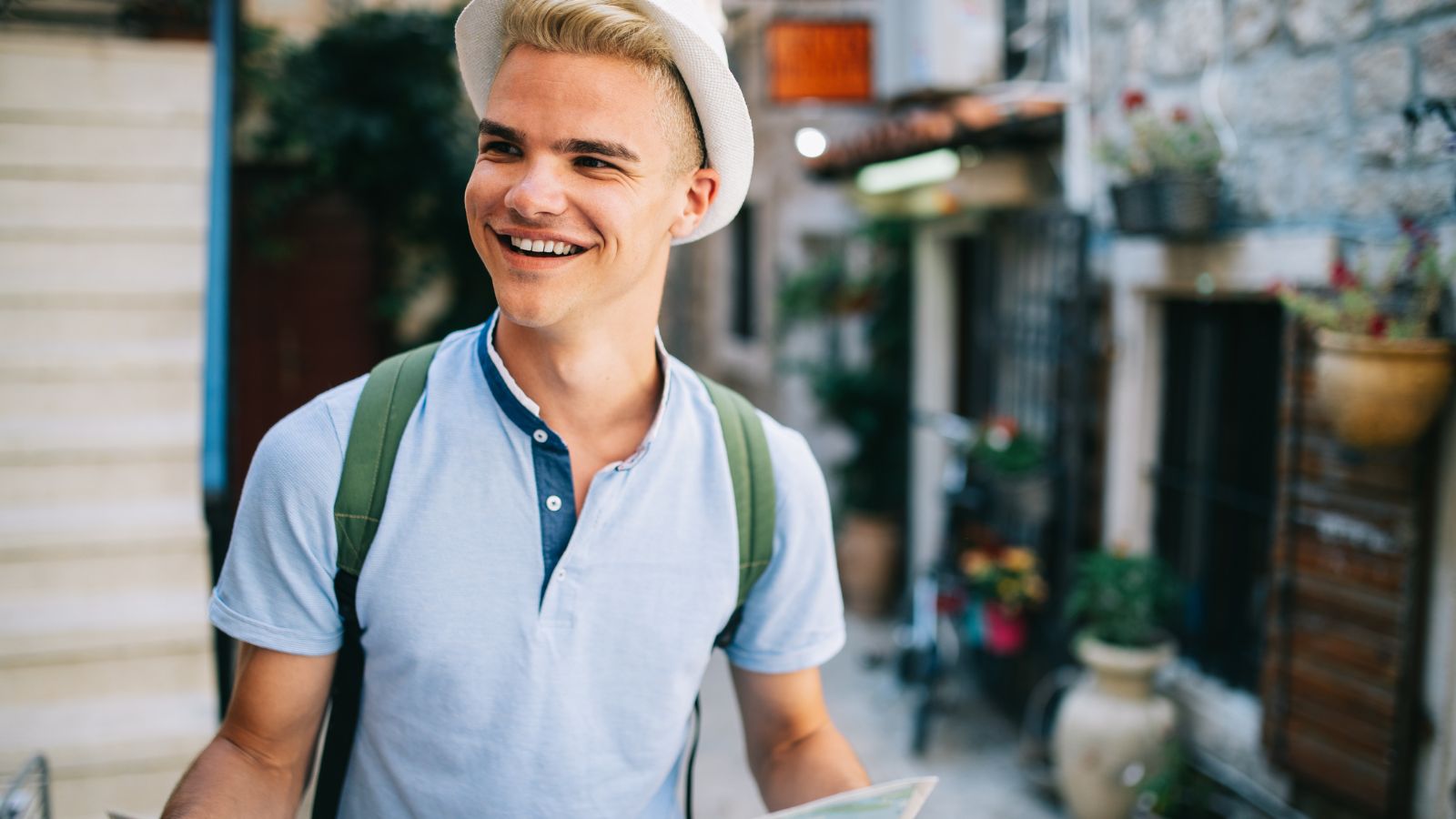 Smiling young man with a hat and backpack holding a map, standing on a sunny street.