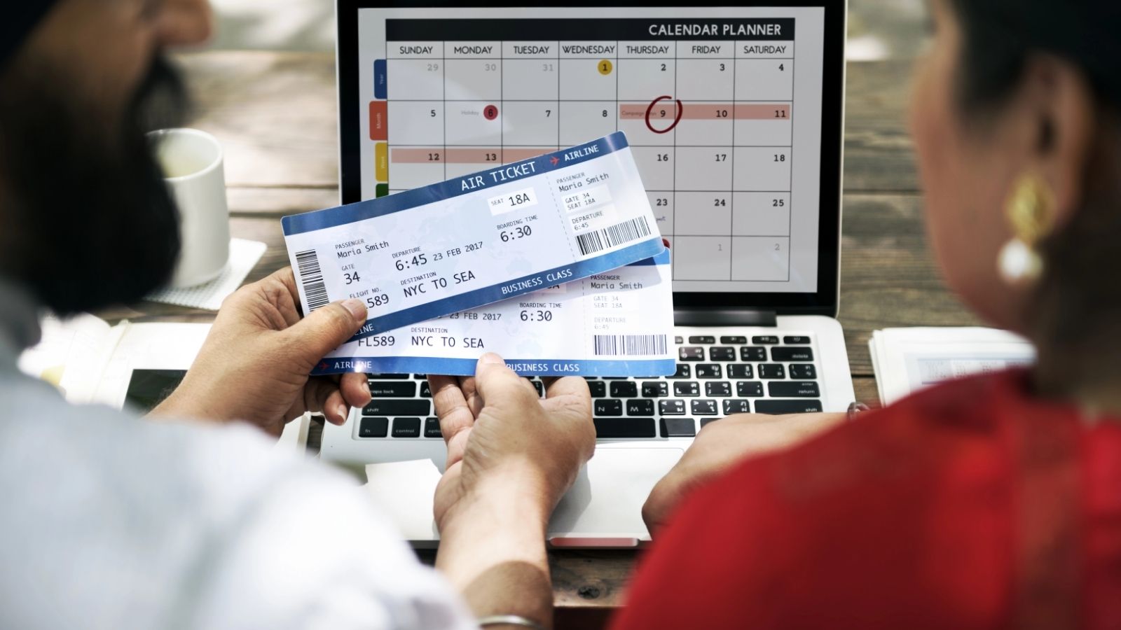 Two people hold airline tickets in front of a laptop displaying a calendar planner.