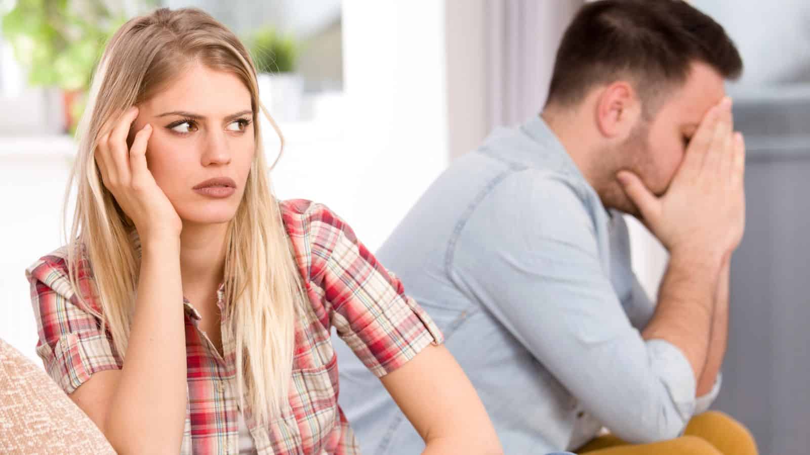 A woman looks frustrated while a man sits behind her with his head in his hands, both appearing upset.