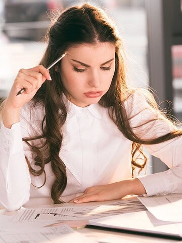 Woman in a white shirt studying documents at a desk with a pen, clipboard, and laptop.