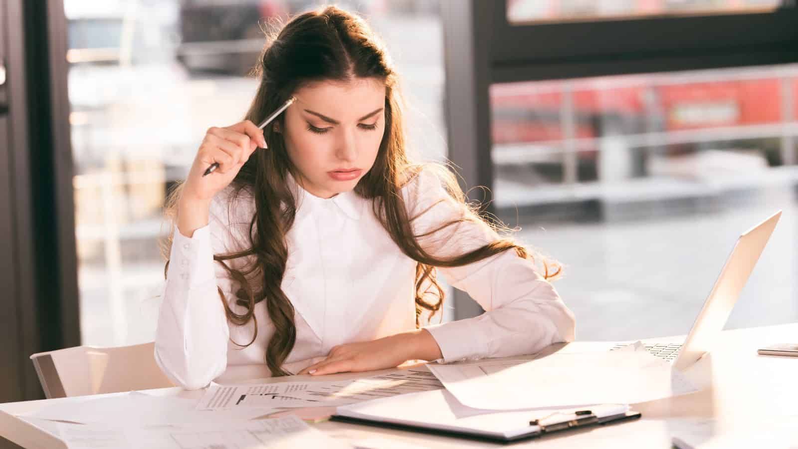 Woman in a white shirt studying documents at a desk with a pen, clipboard, and laptop.