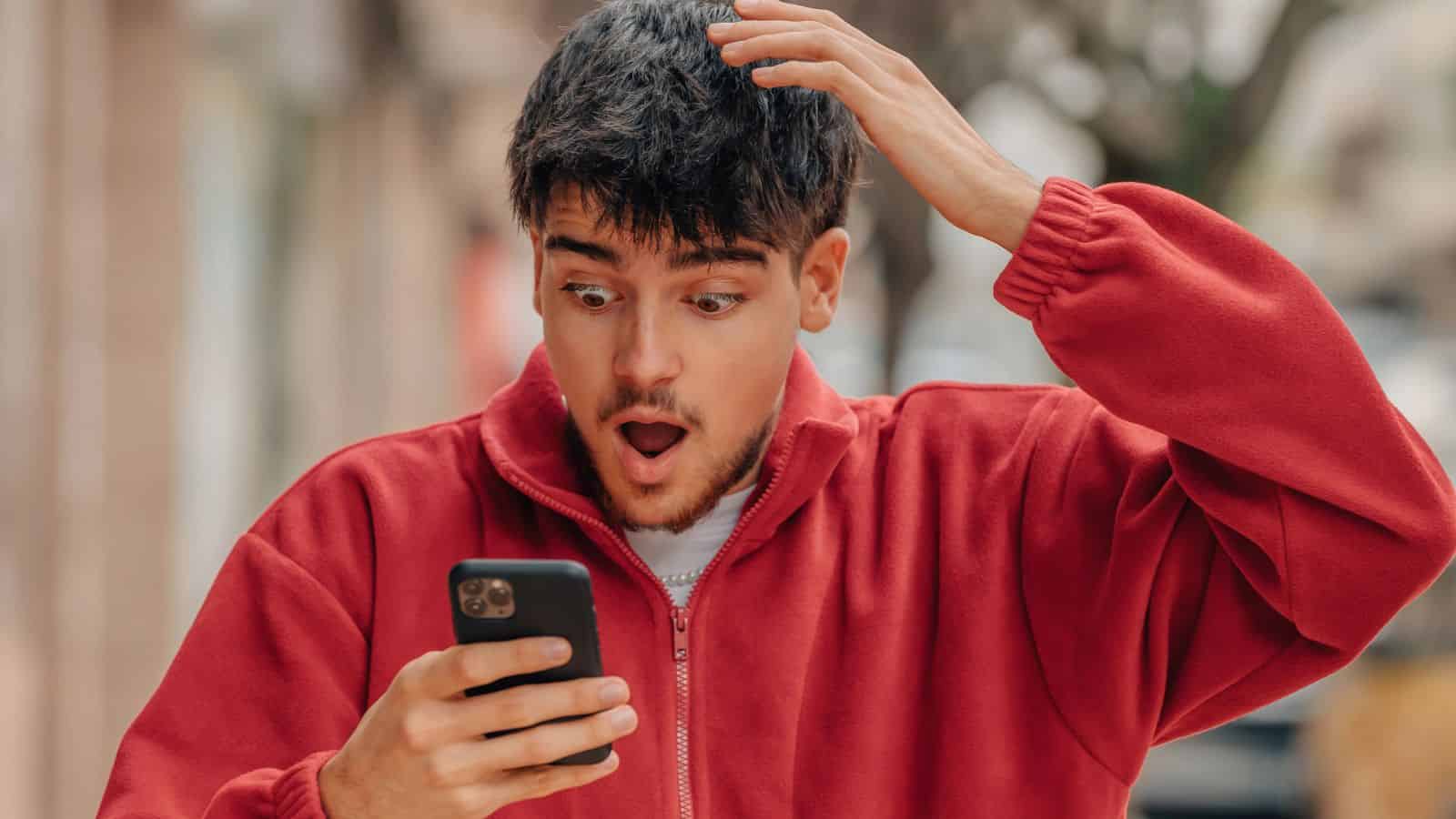 Man in a red jacket looks surprised while holding a phone and touching his head outdoors.