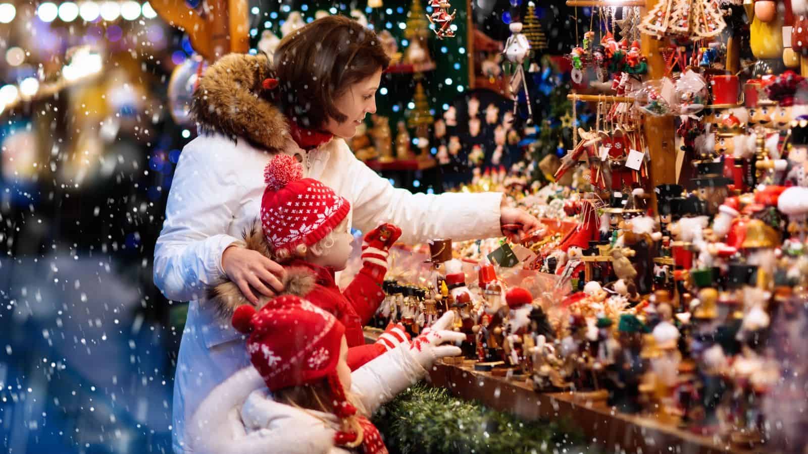 A woman and two children in winter clothes look at toys at a festive outdoor market in the snow.