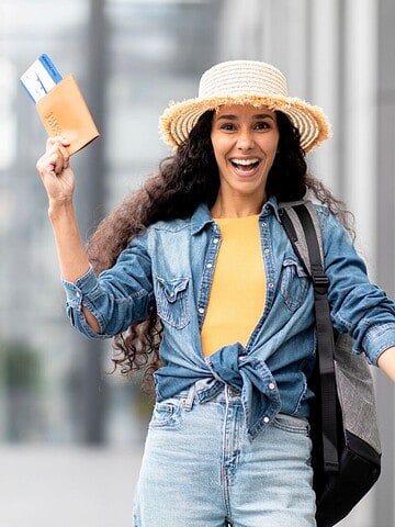 Smiling woman in a hat holding passport and tickets, pulling suitcase outdoors, ready to travel.