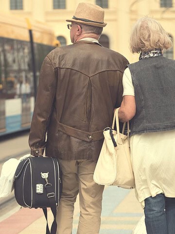 Older couple with suitcases walking at a tram station, with a yellow tram on the tracks nearby.