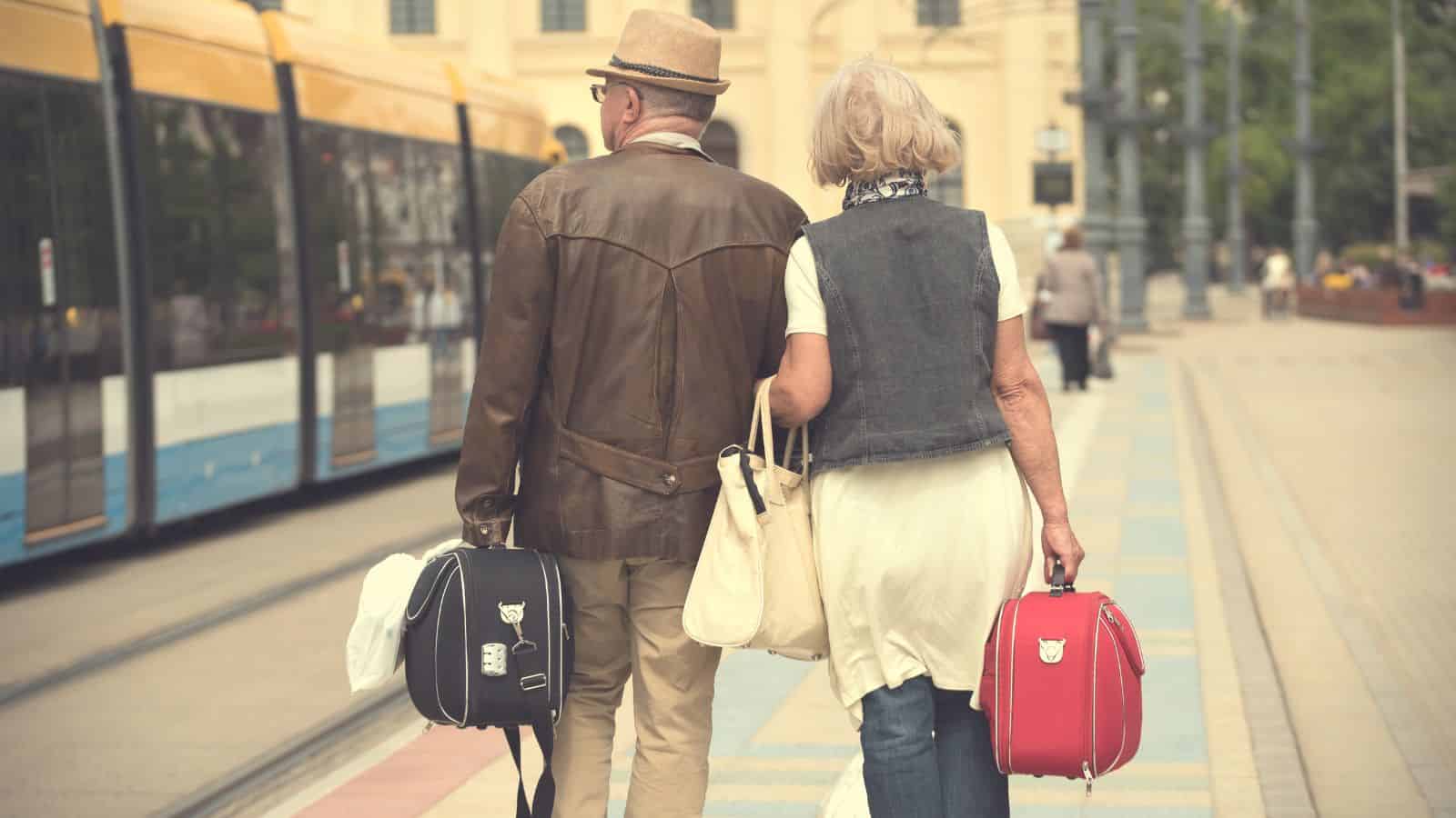 Older couple with suitcases walking at a tram station, with a yellow tram on the tracks nearby.