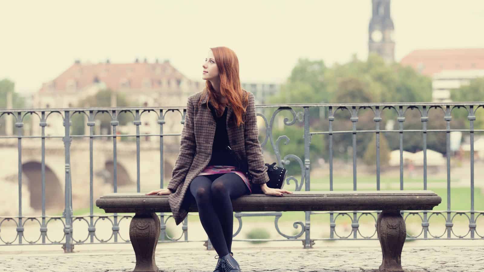 Woman with red hair sitting on a bench by a railing, looking to the side, cityscape in the background.
