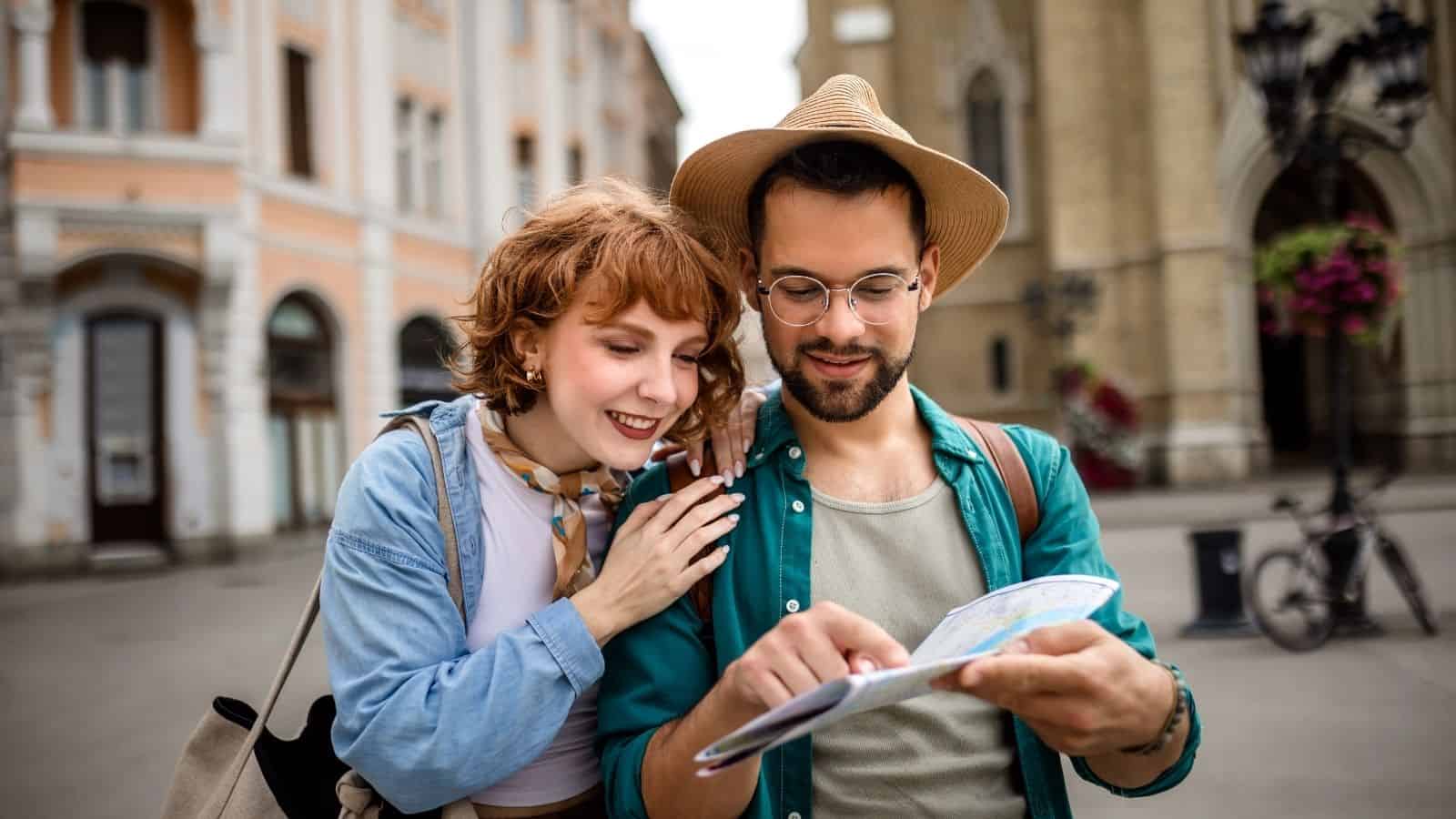 A smiling couple stands outdoors, reading a map together in a city with historic buildings.