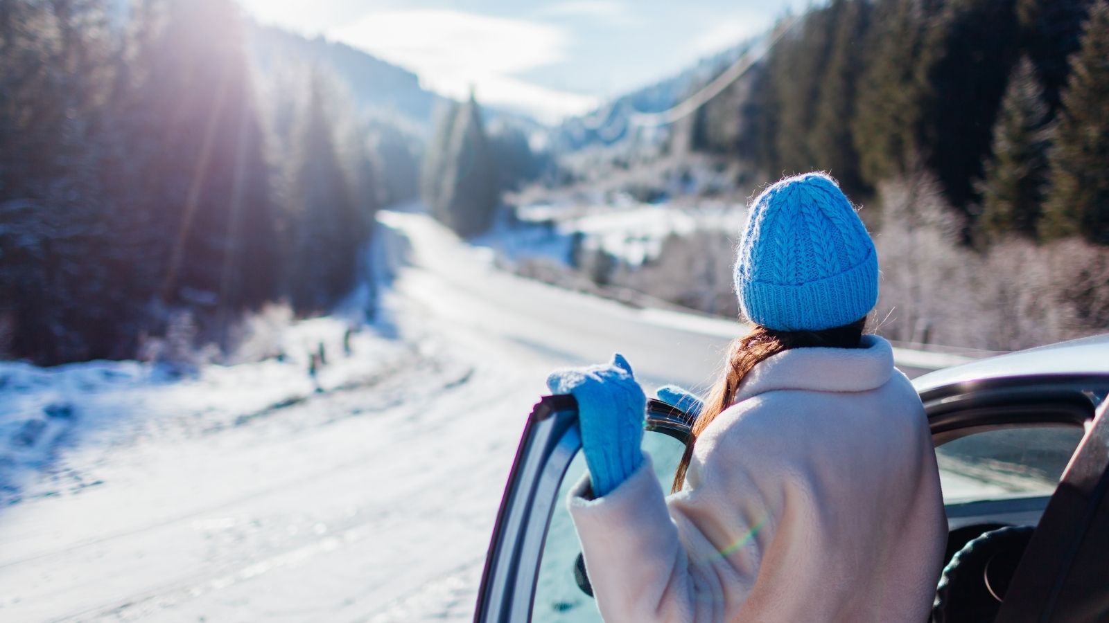 A person in a blue hat stands by a car, looking at a snowy mountain road lined with trees.
