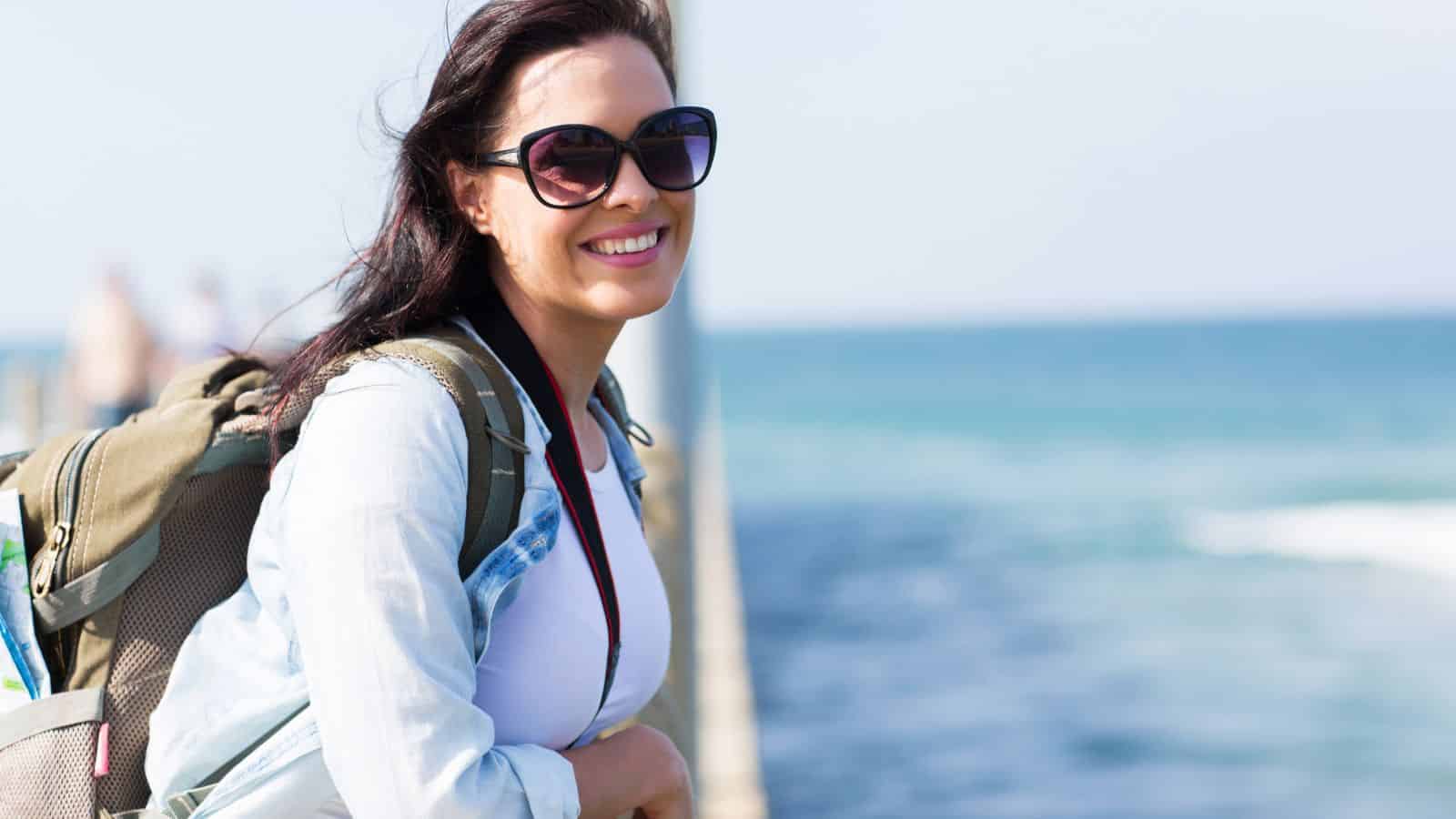Woman with sunglasses and a backpack smiling by the ocean on a sunny day.