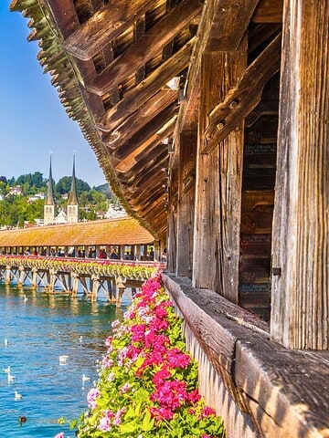 Wooden bridge with flowers crosses a river in a European city with historic buildings under a clear blue sky.