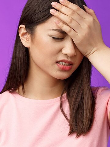Woman in a pink shirt holding her forehead, looking frustrated, against a purple background.