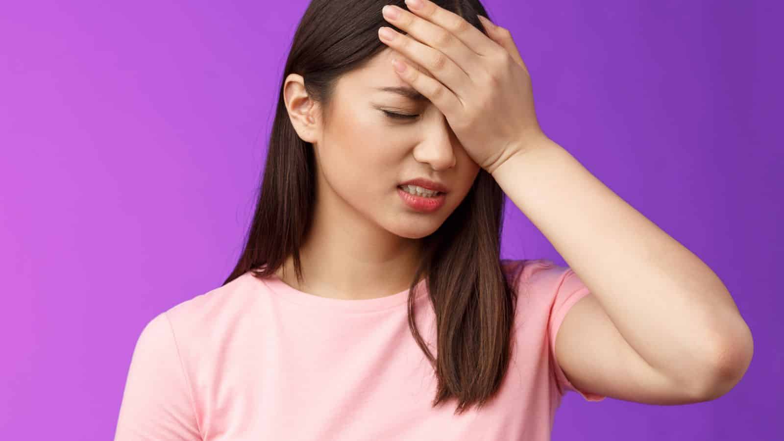 Woman in a pink shirt holding her forehead, looking frustrated, against a purple background.