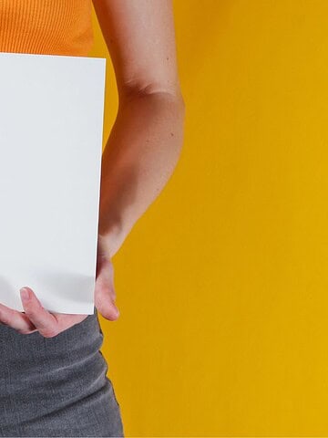 Person in an orange top holding a blank white sheet of paper against a yellow background.