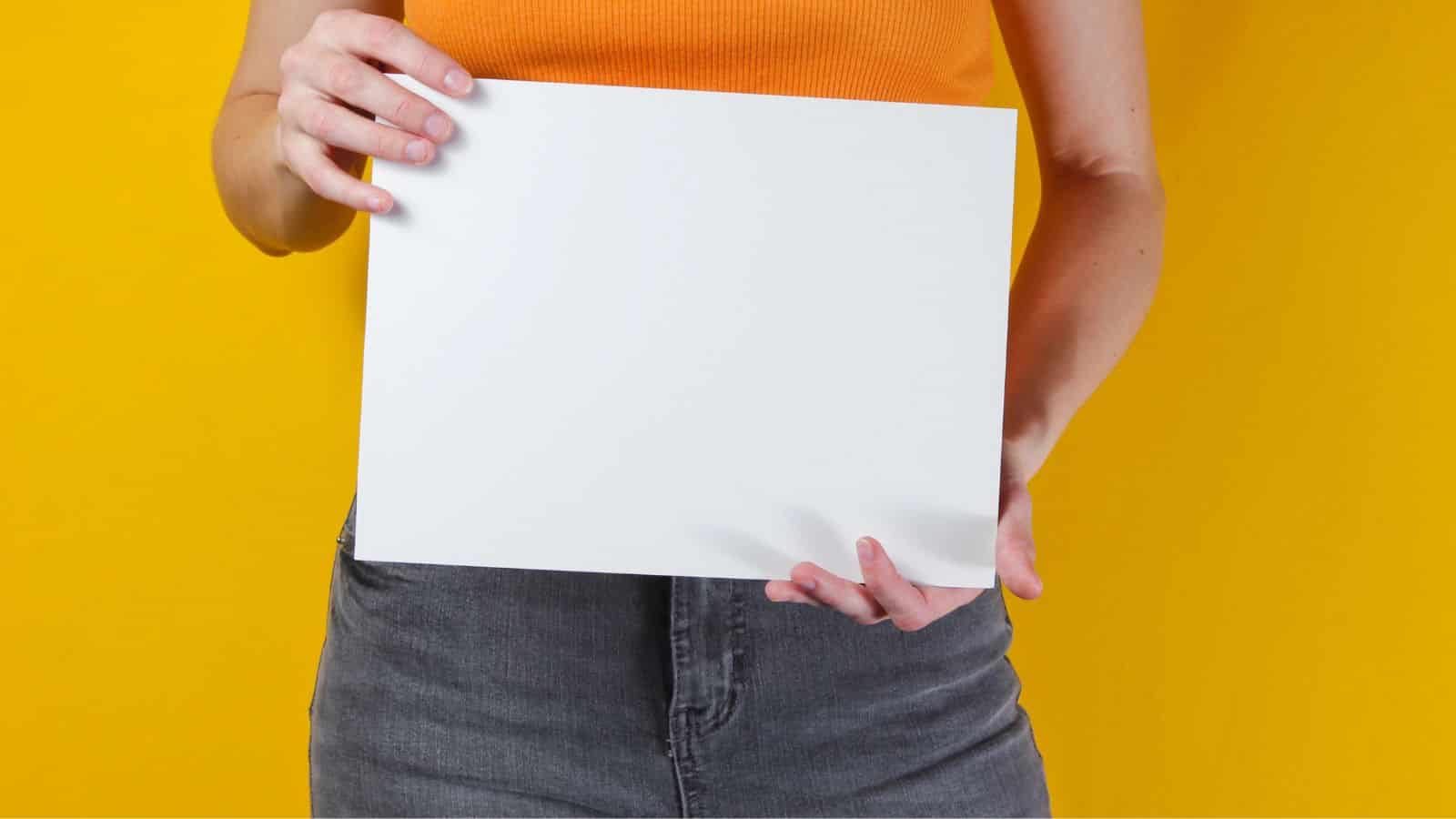 Person in an orange top holding a blank white sheet of paper against a yellow background.