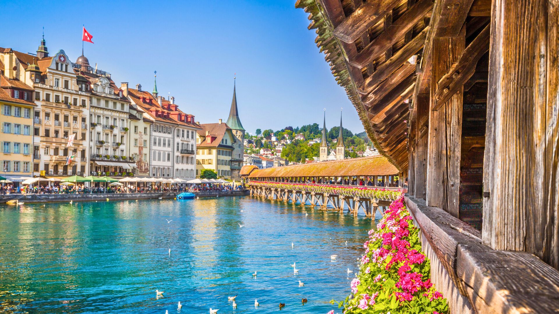 Wooden bridge with flowers crosses a river in a European city with historic buildings under a clear blue sky.
