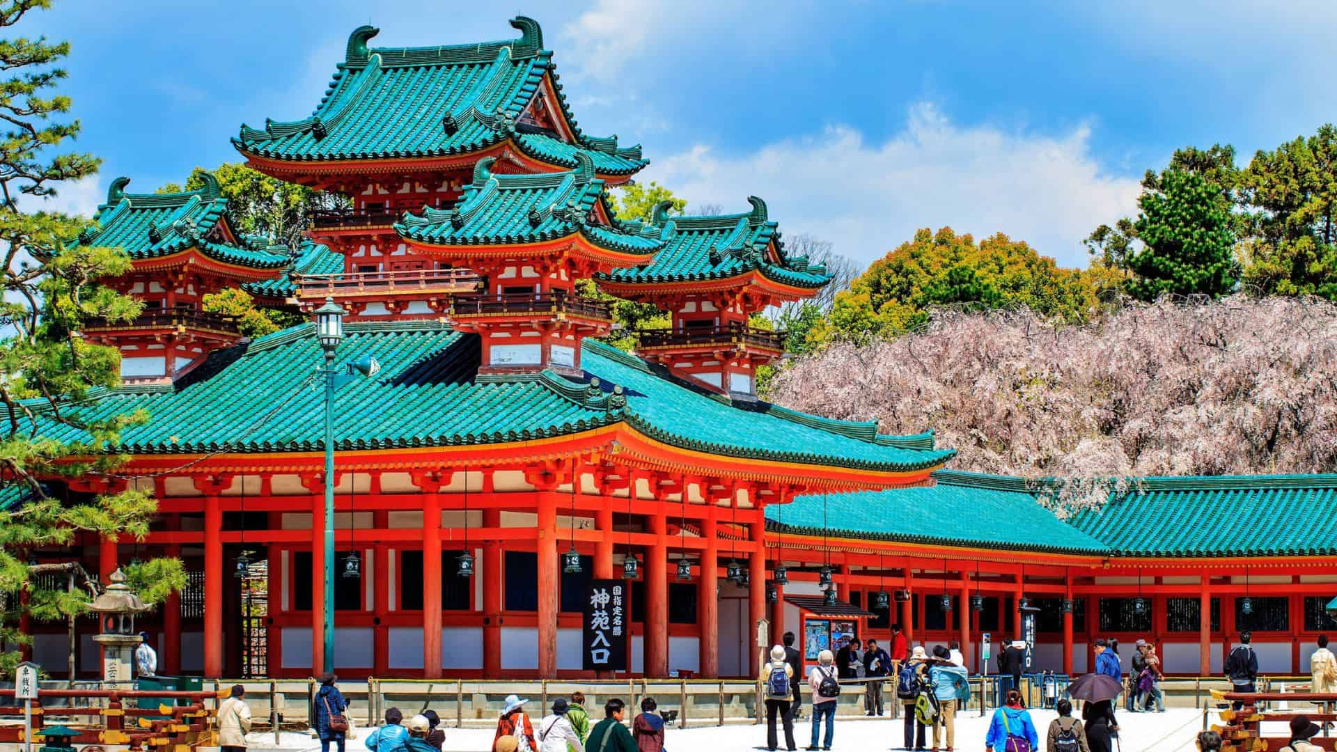 A vibrant red and green Japanese temple with cherry blossoms and people walking in front under a blue sky.