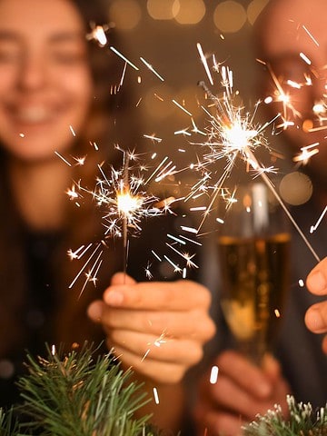 Three people holding sparklers and champagne glasses, celebrating together with festive lights in the background.
