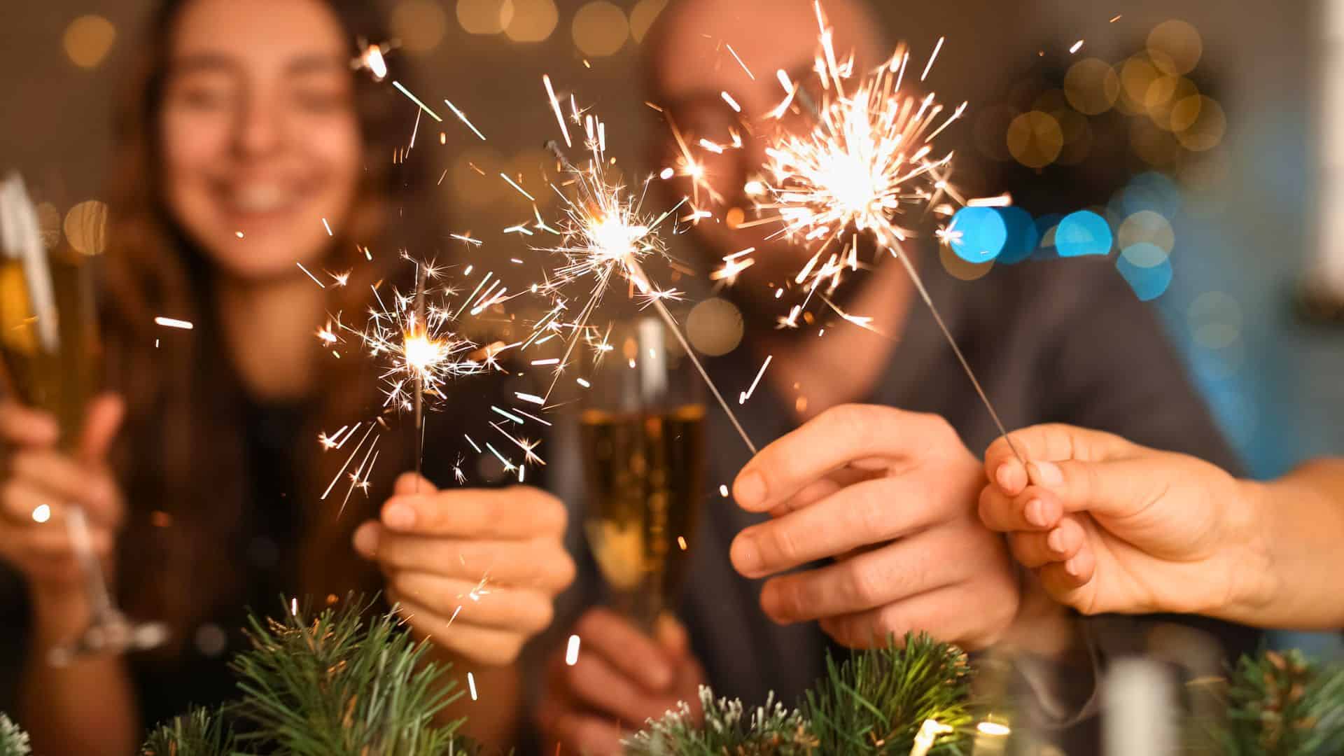 Three people holding sparklers and champagne glasses, celebrating together with festive lights in the background.