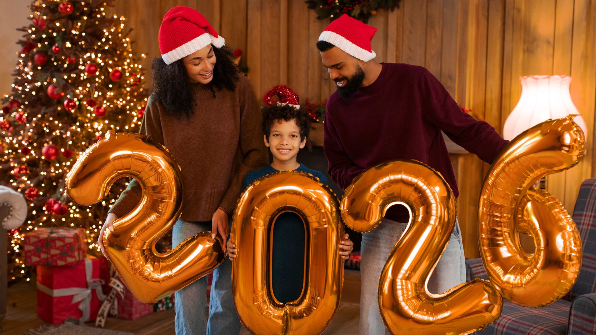 A smiling family in Santa hats holds gold 2026 balloons by a Christmas tree and festive decorations.