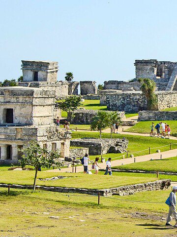 Tourists explore ancient Mayan ruins in Tulum, Mexico, surrounded by green lawns and palm trees.
