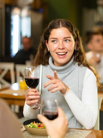 Smiling woman holding a glass of red wine and talking at a restaurant table, with people in the background.