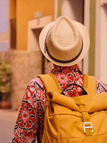 Person wearing a hat and floral shirt with a yellow backpack walking down a colorful narrow street.