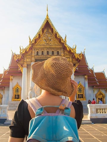 A traveler with a backpack and hat stands facing a golden Thai temple under a bright sky.