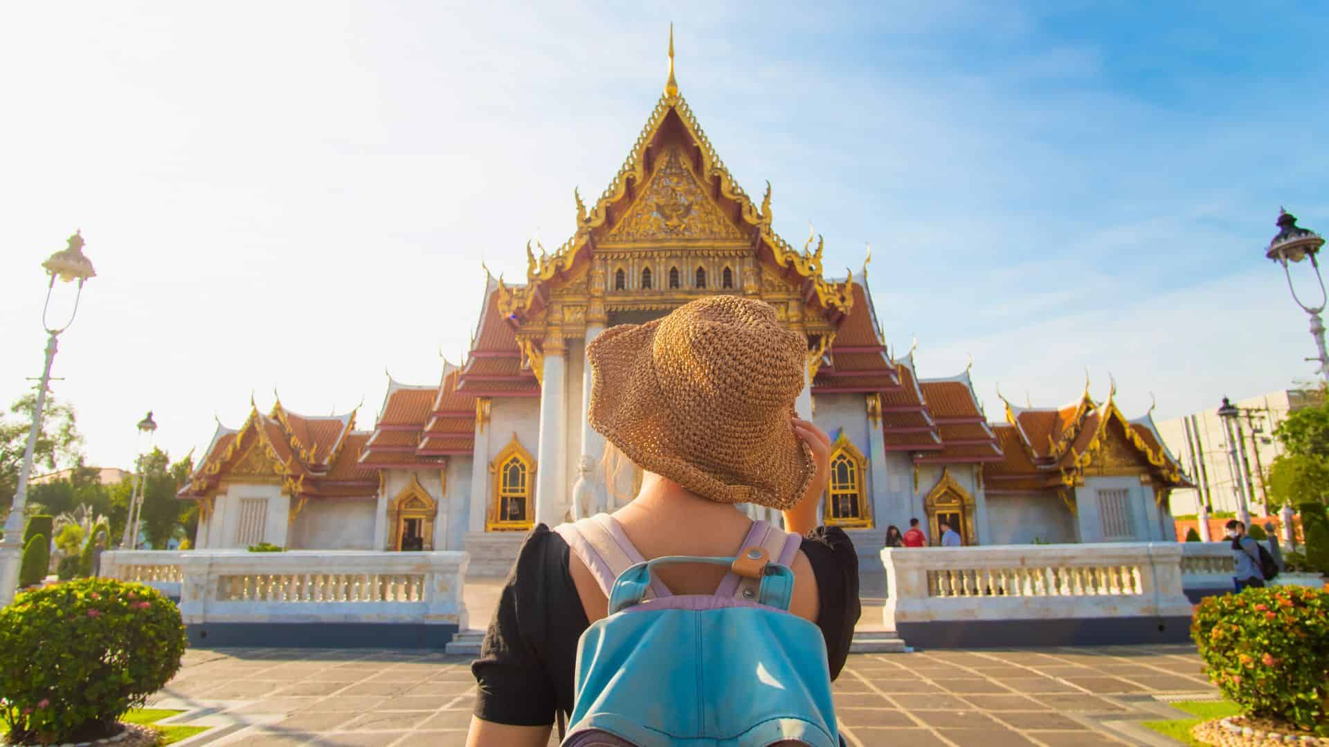 A traveler with a backpack and hat stands facing a golden Thai temple under a bright sky.