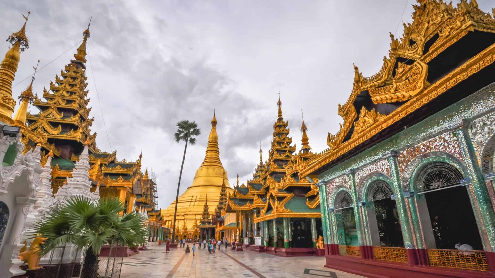 Golden Shwedagon Pagoda with ornate buildings and visitors under a cloudy sky in Yangon, Myanmar.