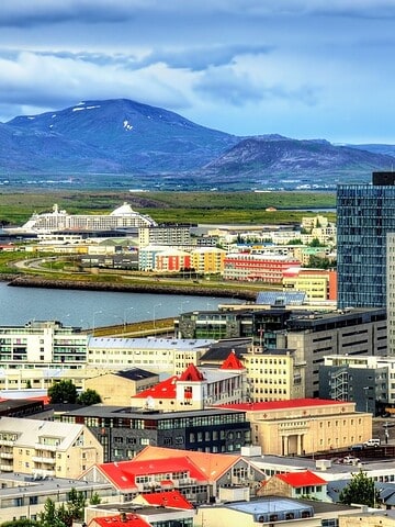 Colorful city buildings near water with mountains in the background under a partly cloudy sky.
