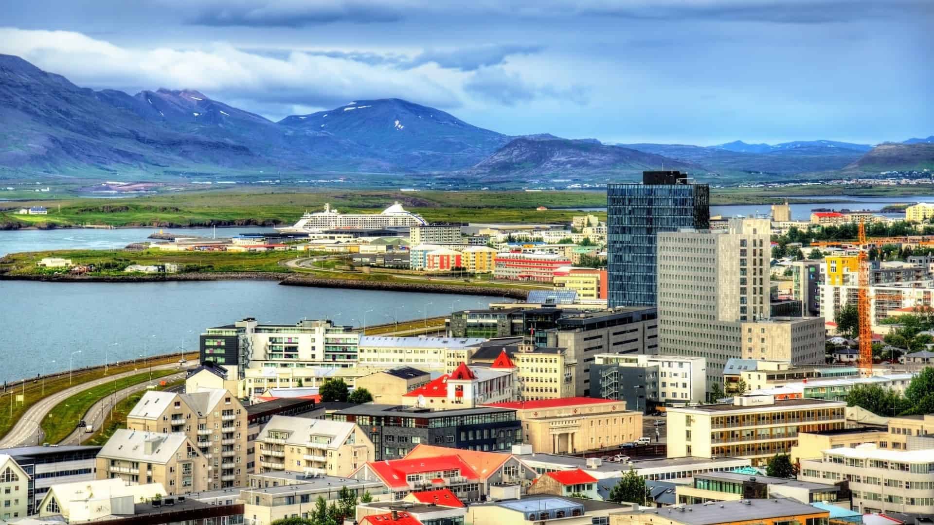 Colorful city buildings near water with mountains in the background under a partly cloudy sky.