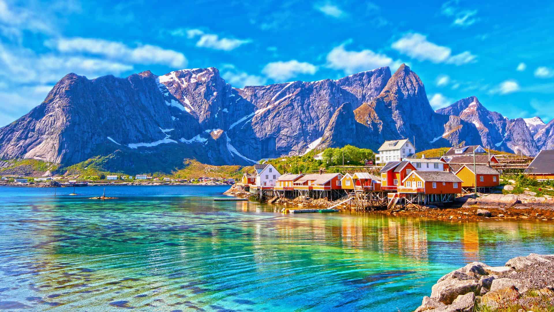Colorful houses on a calm fjord with towering mountains in the background under a bright blue sky.