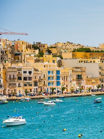Colorful buildings and boats line a bright blue harbor under a clear sky with a large domed church in the background.