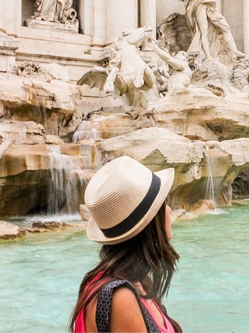 A woman in a hat faces the Trevi Fountain’s flowing water and sculptures in Rome, Italy.