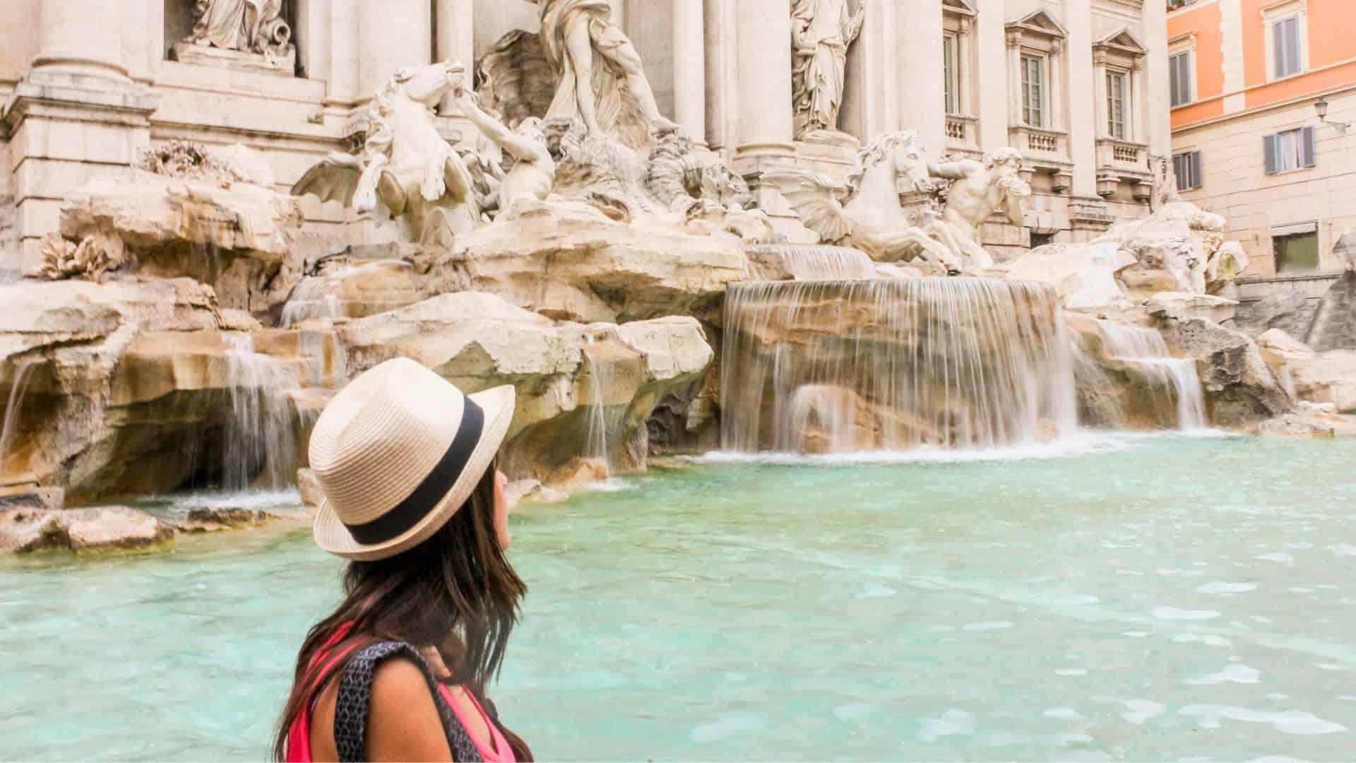 A woman in a hat faces the Trevi Fountain’s flowing water and sculptures in Rome, Italy.