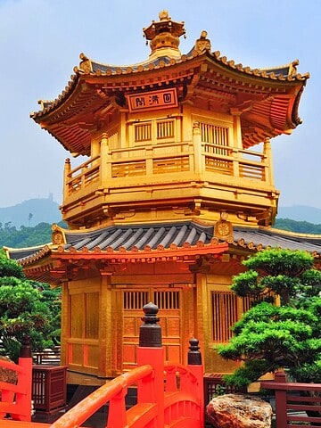A golden pagoda with a red bridge in a lush garden, city buildings and mountains in the background.