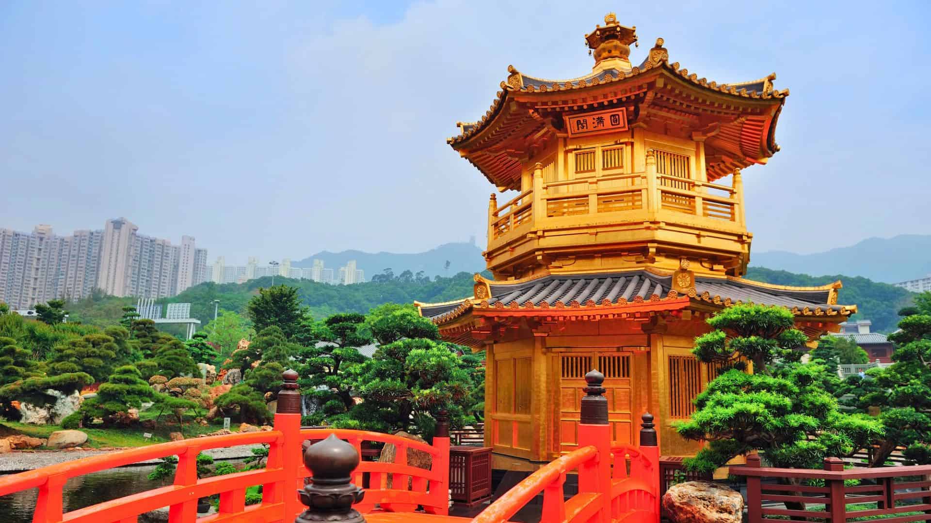 A golden pagoda with a red bridge in a lush garden, city buildings and mountains in the background.