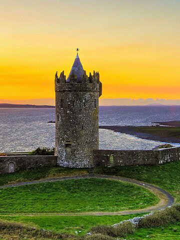 Stone tower sits on green hill overlooking the sea at sunset, with orange and yellow sky in the background.
