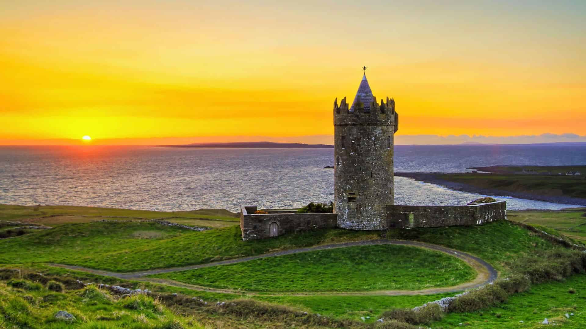 Stone tower sits on green hill overlooking the sea at sunset, with orange and yellow sky in the background.