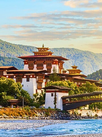 Traditional Bhutanese monastery by a river, surrounded by green hills and a wooden bridge, at sunset.