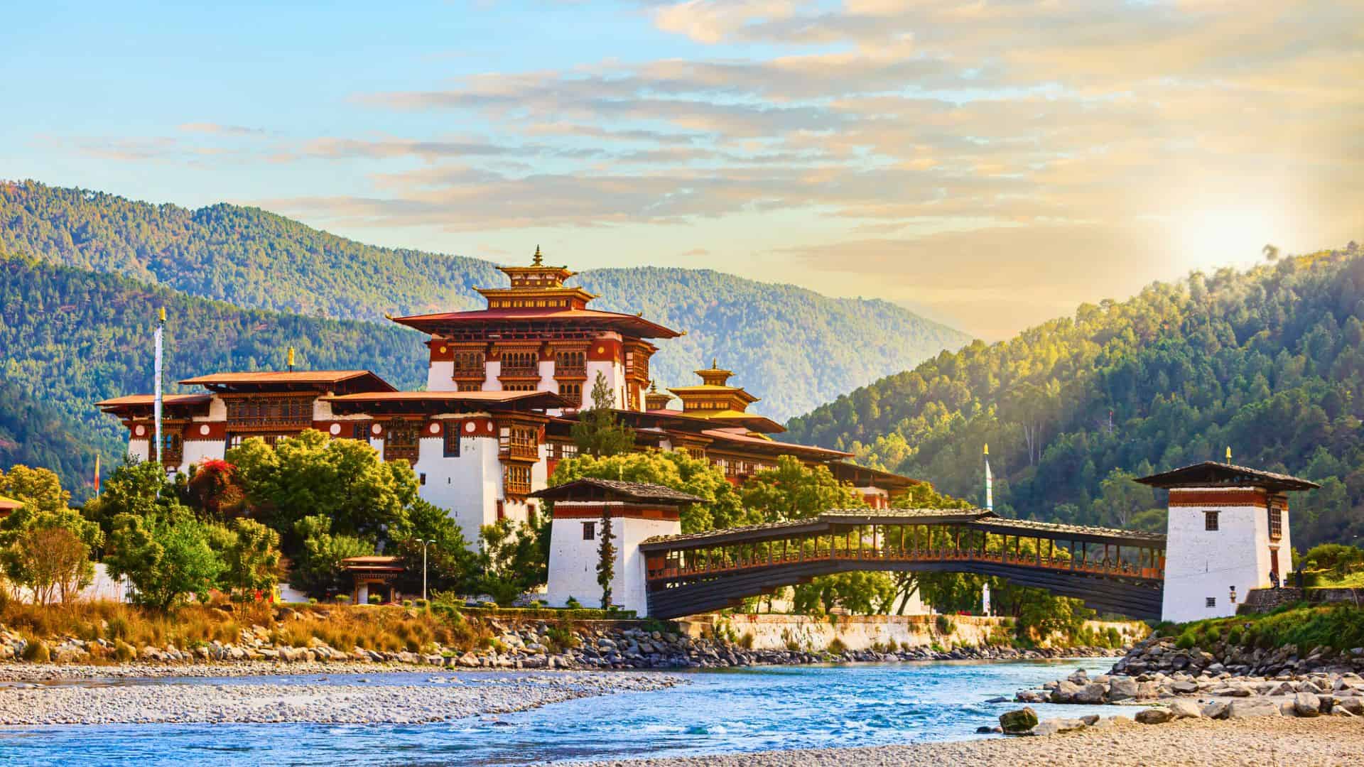 Traditional Bhutanese monastery by a river, surrounded by green hills and a wooden bridge, at sunset.