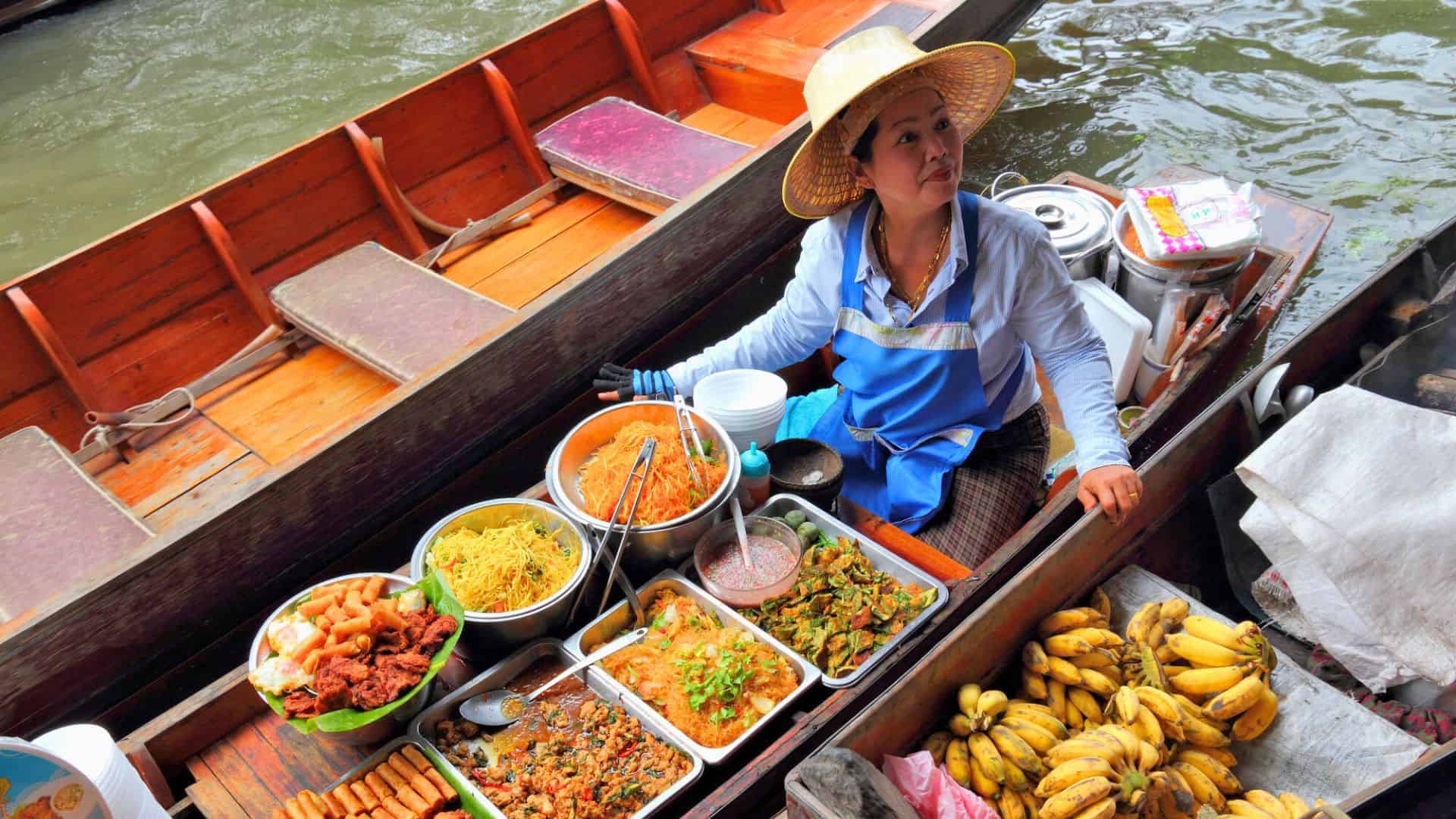 A woman in a straw hat sells various foods and bananas from a boat at a floating market.