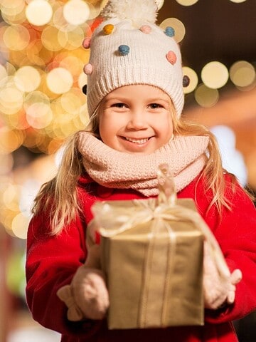 Smiling child in a red coat holds a wrapped gift at a festive, brightly lit outdoor holiday market, surrounded by sparkling holiday lights.