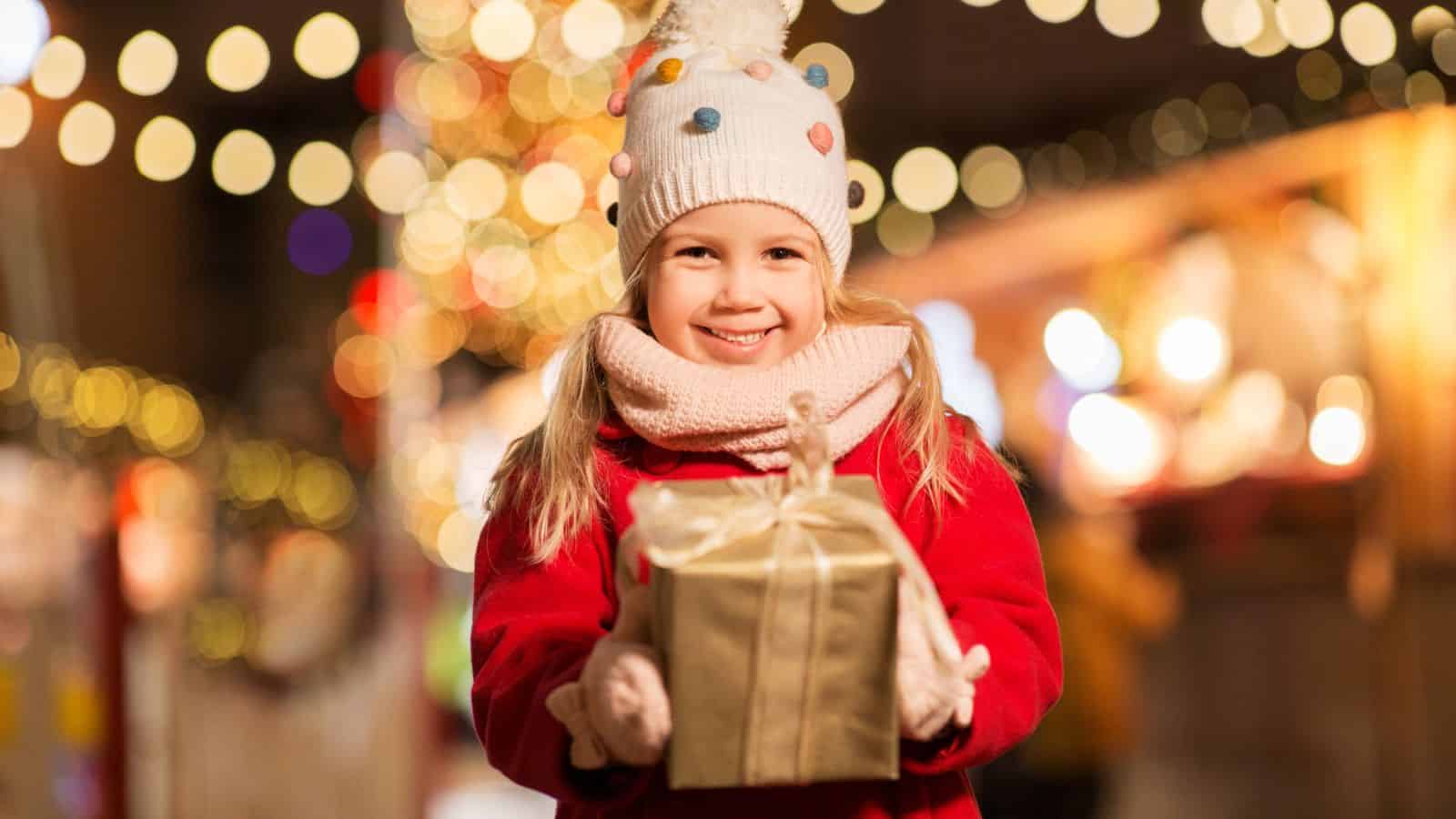 Smiling child in a red coat holds a wrapped gift at a festive, brightly lit outdoor holiday market, surrounded by sparkling holiday lights.