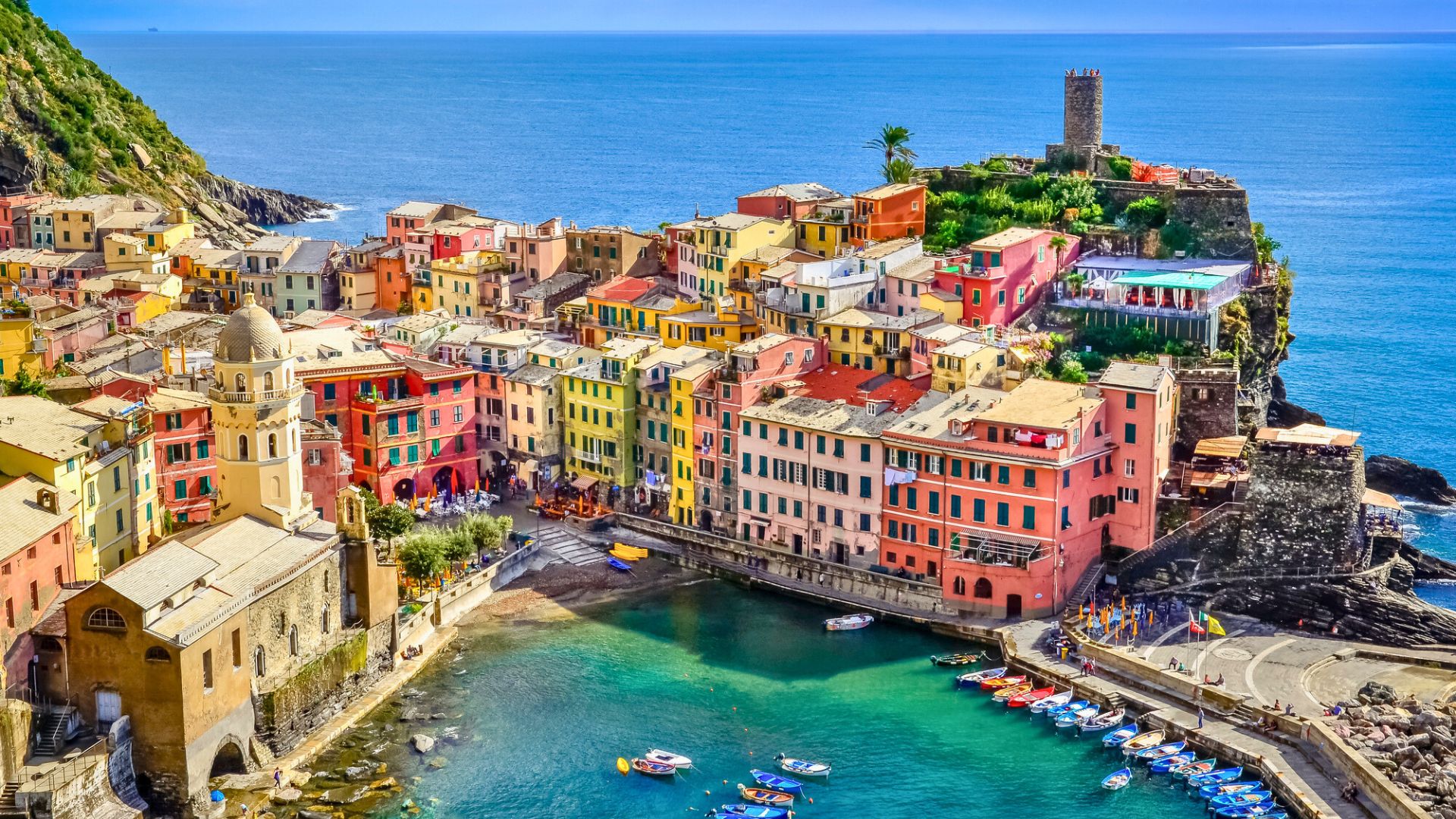 Colorful seaside village with boats in a small harbor, vibrant buildings, and blue sea under a sunny sky.